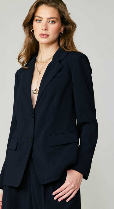 A woman wears a singe breasted navy blazer. There are two buttons that match the fabric. There is a low pocket on each side and a high pocket on the left side.