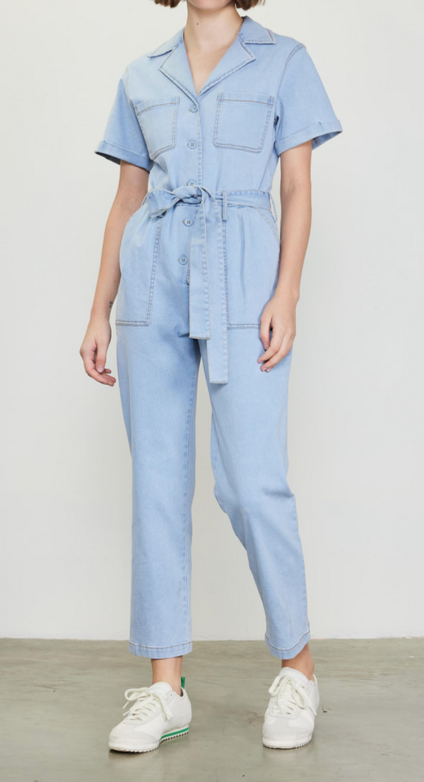 A woman is standing, wearing a blue chambray jumpsuit with short sleeves, a collar, and front pockets. She has a belt tied around her waist and is posing against a plain background.