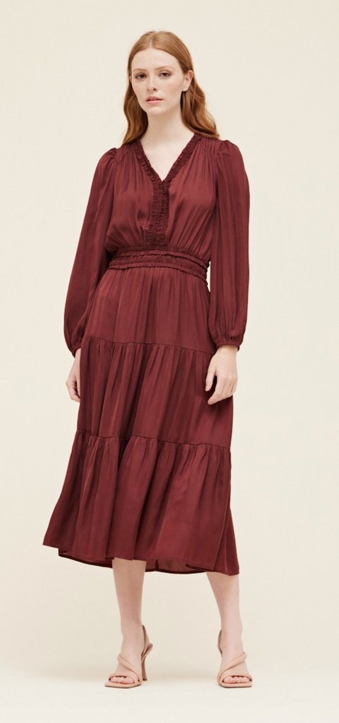 This model is wearing a long sleeve midi dress in a wine color. The skirt is tiered. The waist is cinched in and the neckline is a v shape.