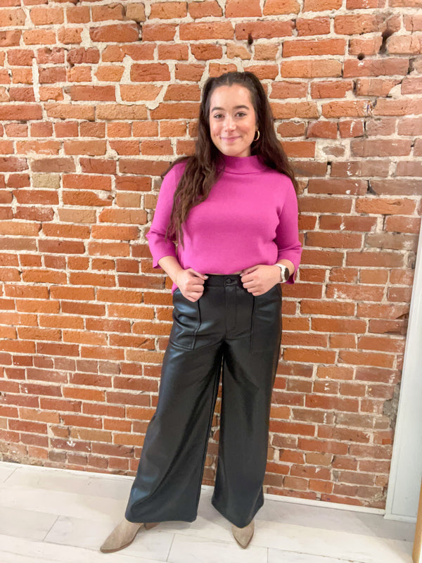 This is a full length picture of a model. She's wearing long wide leg pants in a black faux leather material. She has it paired with a bright pink mock neck top.
