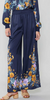 This picture is of a model from the waist down. She is wearing long wide legged pants with a smocked waist. The pants are navy and have a floral print at the hem that runs up the outer side of both legs. The flowers are in goldm light purple, and light blue tones.