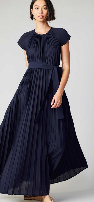 This is a full length picture of a model. She is wearing a short sleeve maxi dress in navy blue. The dress is fully pleated in the bodice and in the skirt. There is a matching belt that ties at the waist. The fabric is slightly silky.