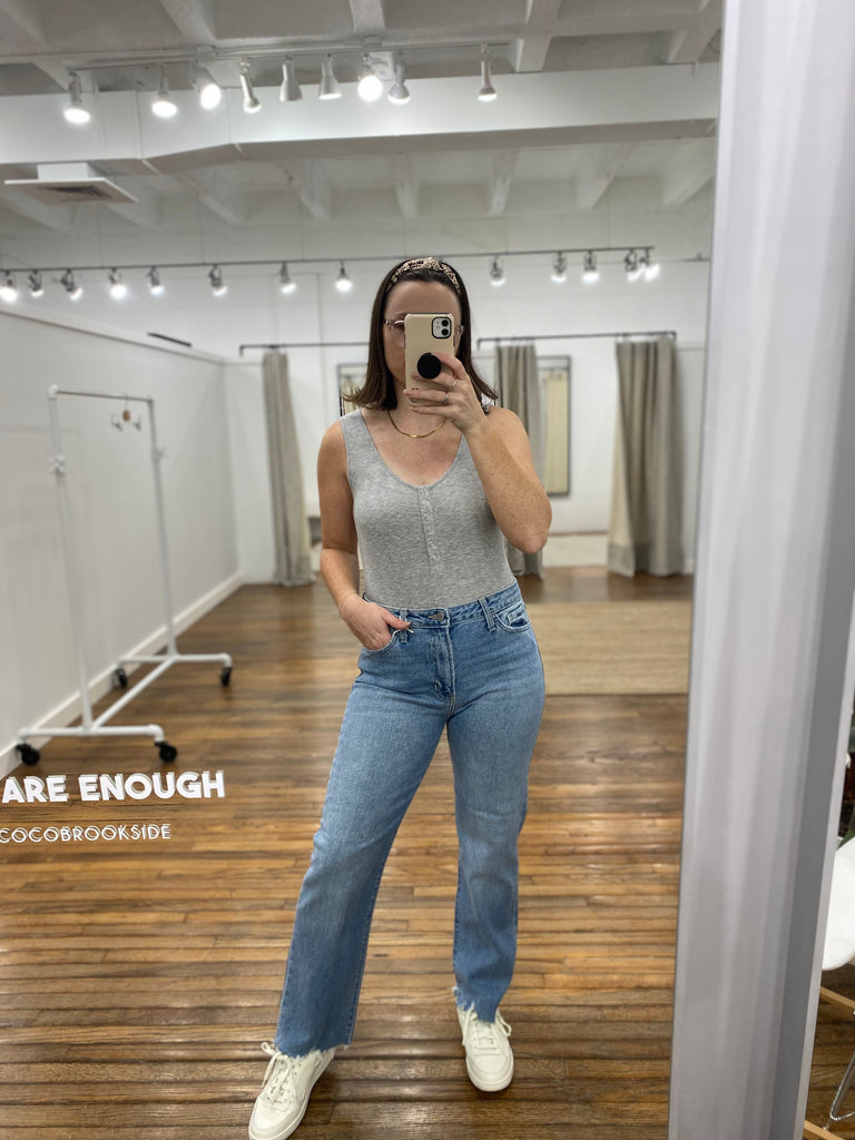This is a mirror selfie of our model. She is wearing a heather grey tank top bodysuit. It has a rounded neckline with six silver buttons down the front. She has it paired with light high waisted denim jeans.
