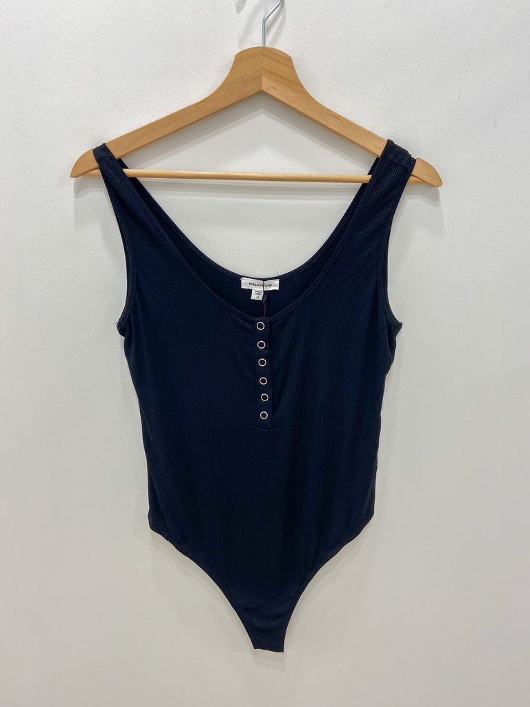 This is a picture of a black bodysuit hanging from a wooden hanger in front of a white wall. The bodysuit is a tank to with a round neckline. There are six silver snaps down the front to open up the neckline.