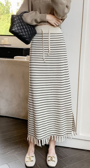 This is a picture of a model from the waist down. She is wearing a knit skirt in ivory with horizontal black stripes. There are frayed tassels along the bottom hem of the skirt. The waist is elastic with draw strings.