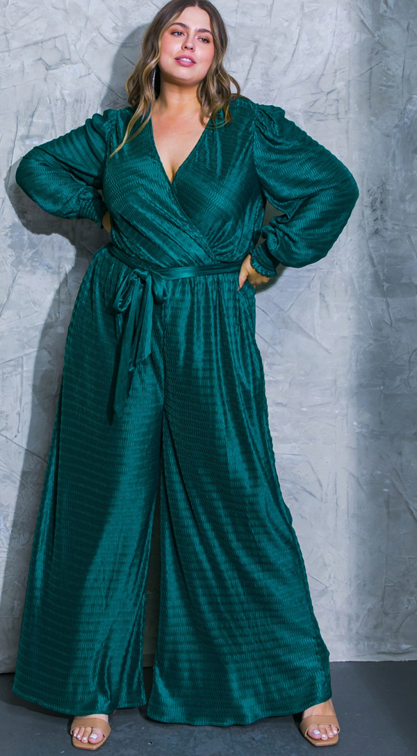 This is a full length picture of a model. She is wearing a long sleeve jumpsuit in teal. The jumpsuit has a deep v neckline. The fabric is textured and there is a matching belt that ties around the waist.