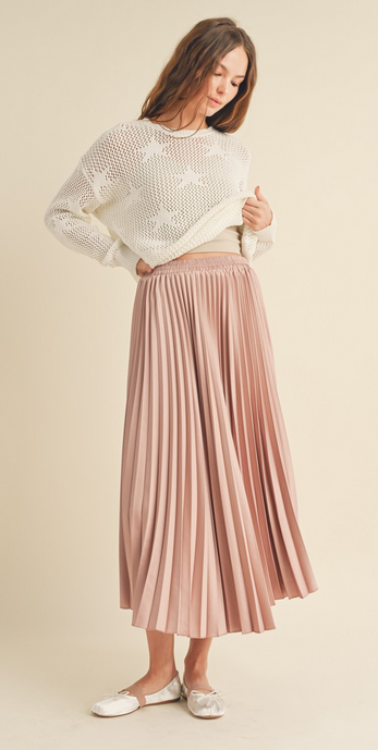This is a full length picture of a model. She's wearing a light pink pleated maxi skirt. The skirt is paired with a white sweater that has white stars on it.