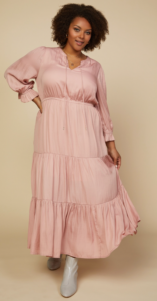 This is a full length picture of a model. She wears a long sleeve light pink maxi dress that comes down to her ankles. The waist of the dress is higher and the skirt is tiered.