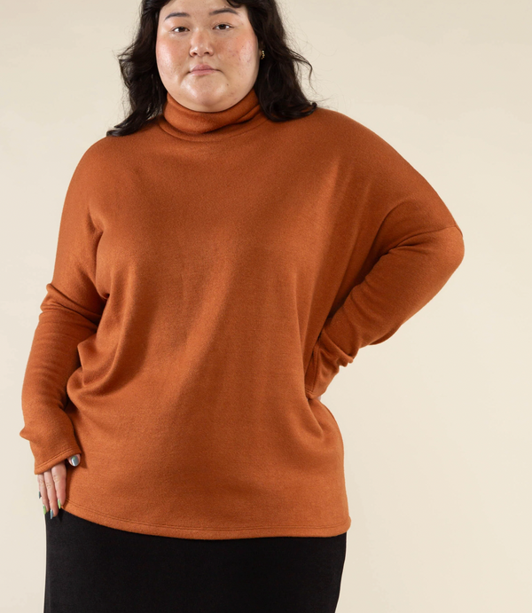 This is a nearly full length picture of a model. She is wearing a light long sleeve turtle neck sweater in a bronze color.