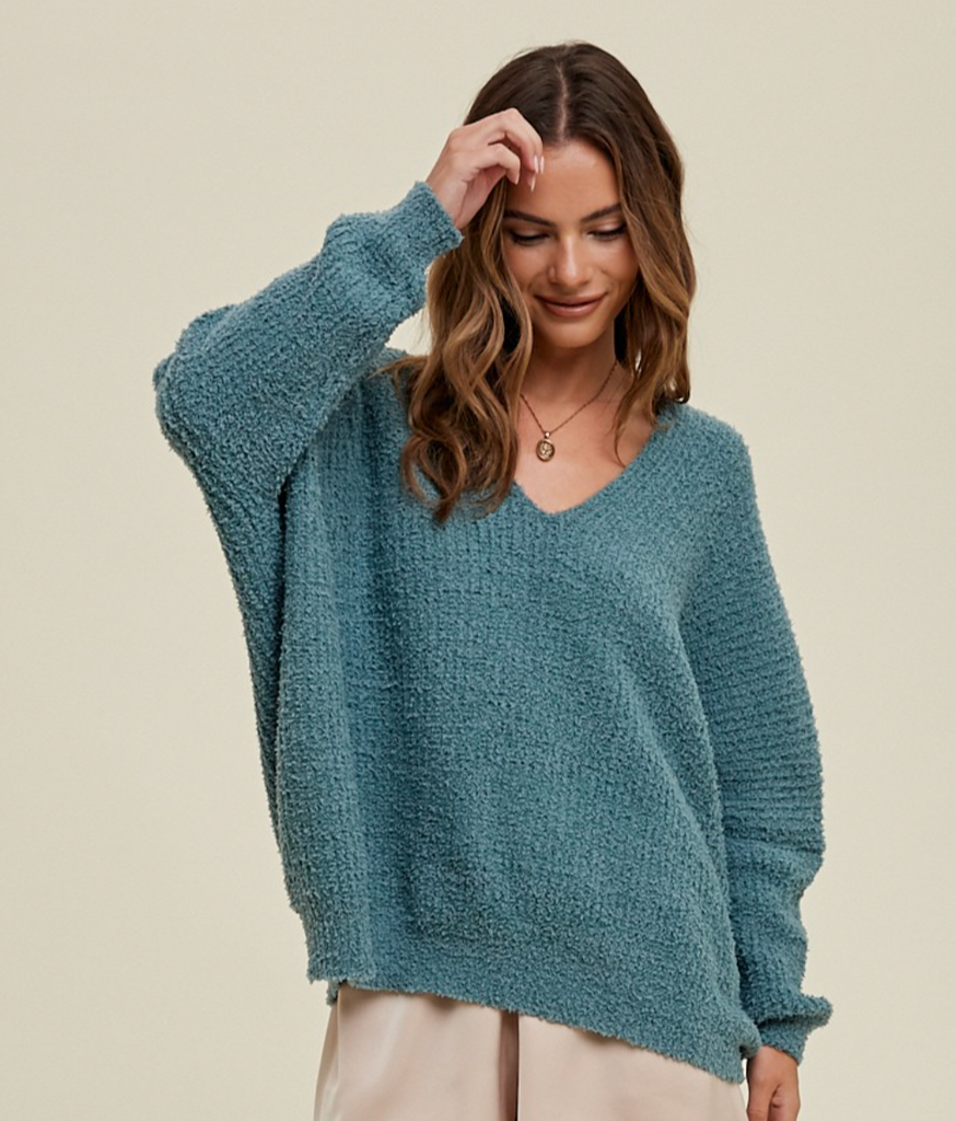 This is a picture of a model from the waist up. She is wearing a very soft oversized long sleeve sweater in a dusty blue. The sweater has a v neckline.