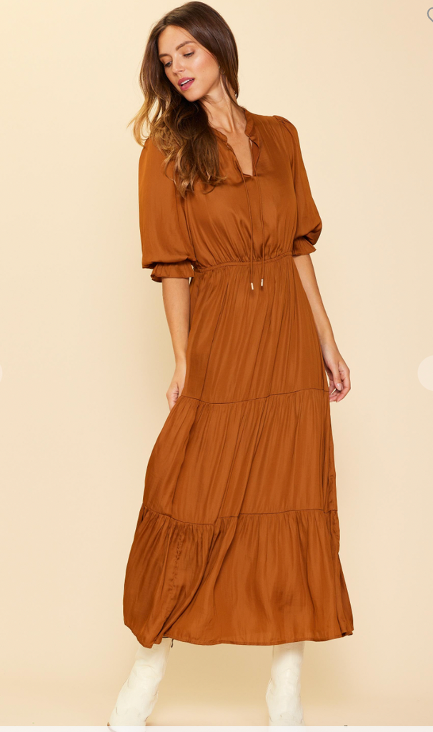 This is a full length picture of a model. She wears a long sleeve caramel colored maxi dress that comes down to her ankles. The waist of the dress is higher and the skirt is tiered.