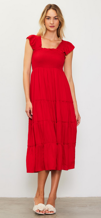 This model is wearing a midi length dress in a bright red. The bodice is smocked and has romatic flutter sleeves. The skirt of the dress is tiered.