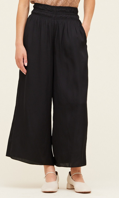 A picture of a model from the waist down. She's wearing black wide leg pants with a smocked waist. The pants are a satin fabric.