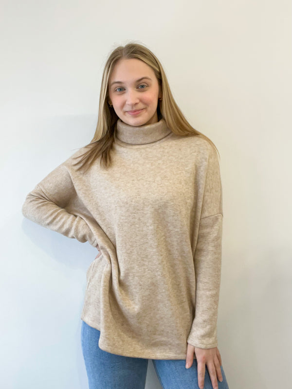 This is a nearly full length picture of a model. She is wearing a light long sleeve turtle neck sweater in an oatmeal color.
